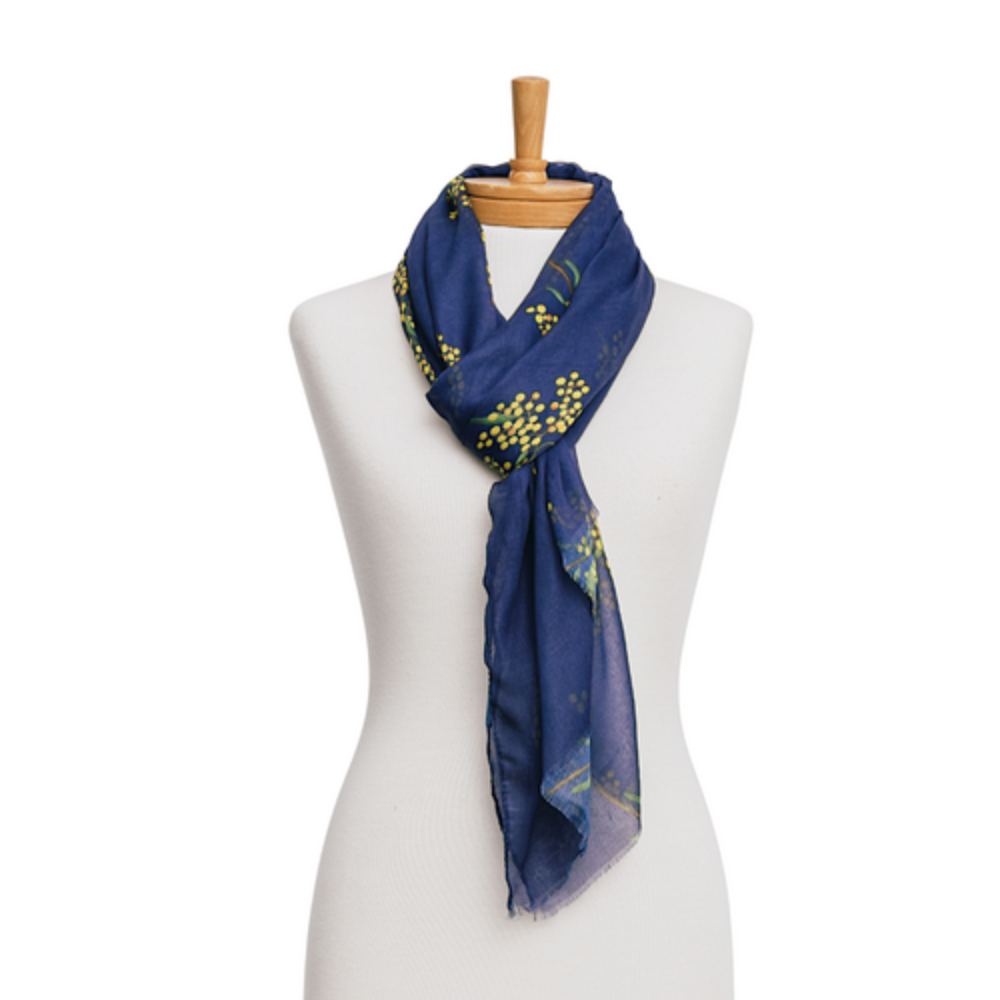 a lightweight cotton scarf that has a navy blue background and bunches of yellow Wattle flowers with green leaves. The scarf is tied around a mannequin's neck.