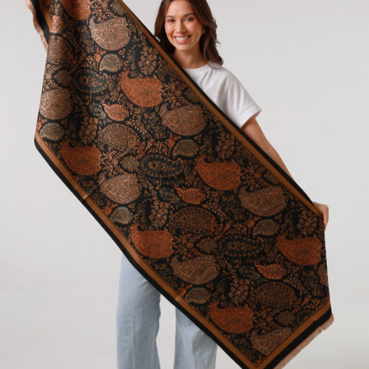 Woman holding a warm and cosy ladies scarf with autumn tones and a large paisley pattern on a grey background.