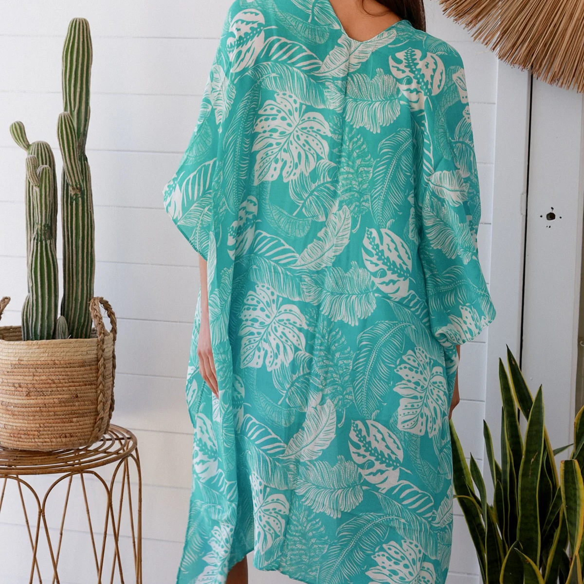 Woman wearing a teal leaf-patterned cover-up standing in a room with plants and a white wall.