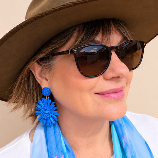 a woman wearing a hat, sunglasses, blue fflower earrings and a blue scarf over a white top.