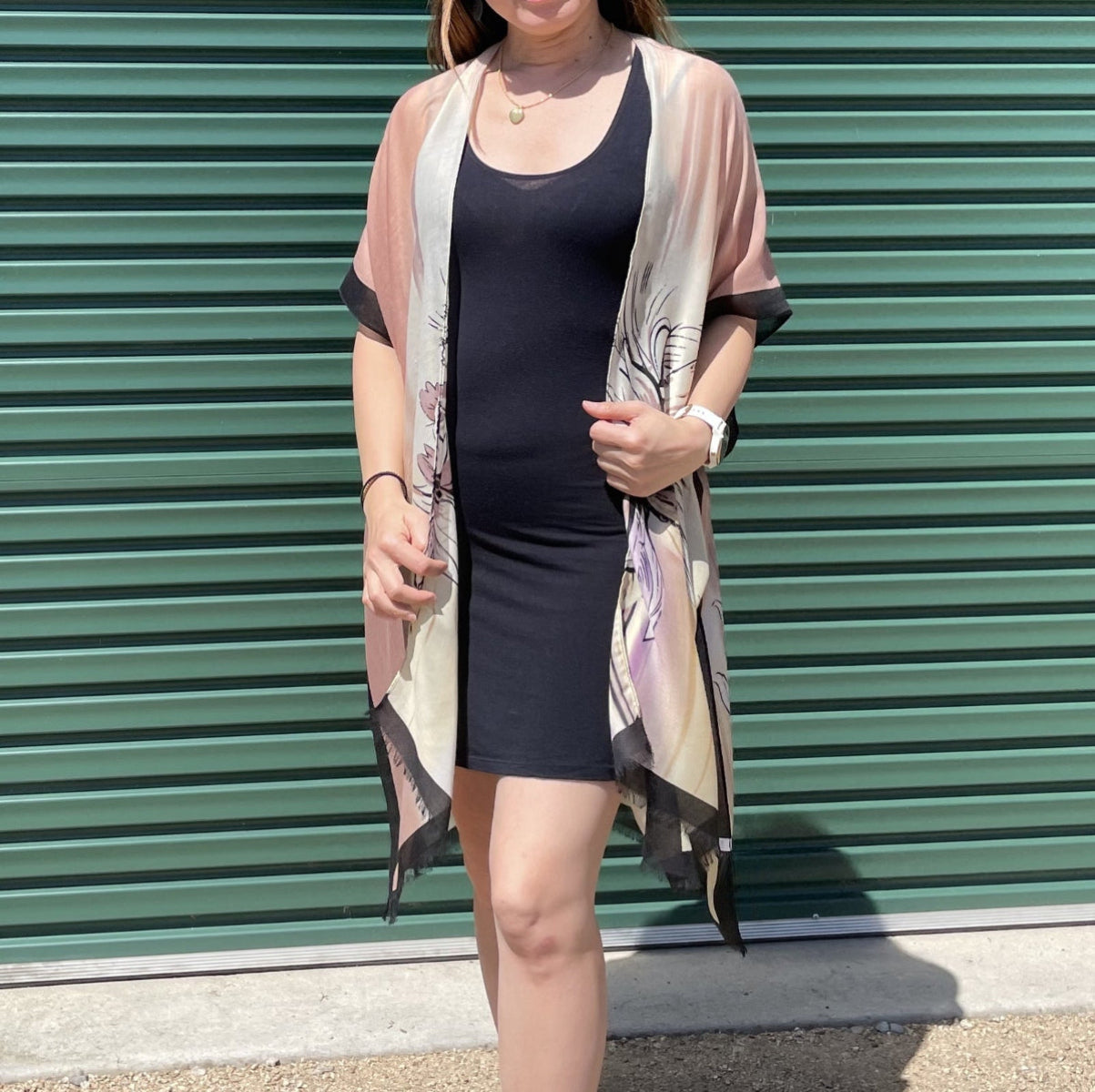Woman wearing a black dress and beige kimono-style jacket against a green metal shutter.