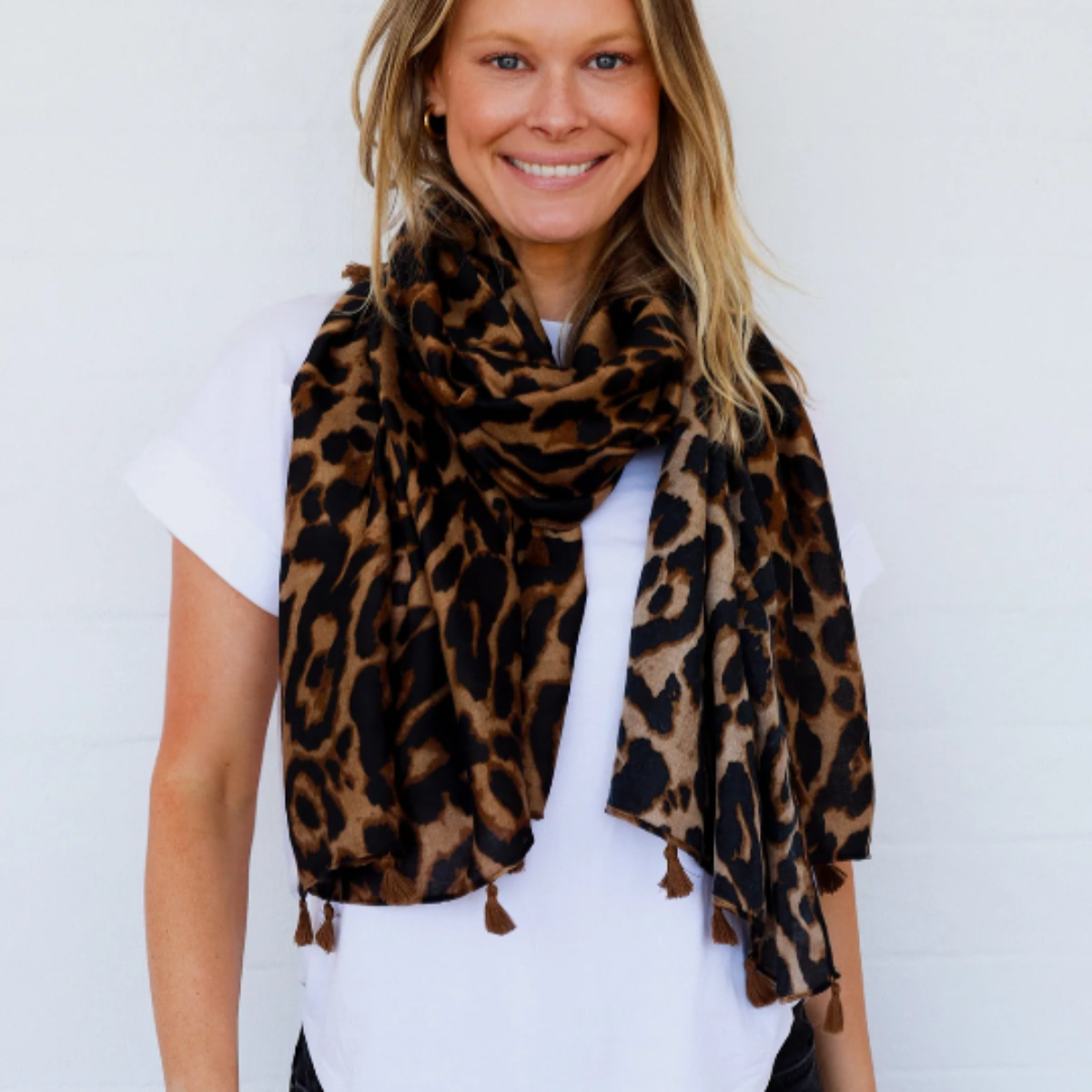 a lady wearing a leopard scarf in colours of caramel brown and black, The scarf is finished with caramel coloured tassels.