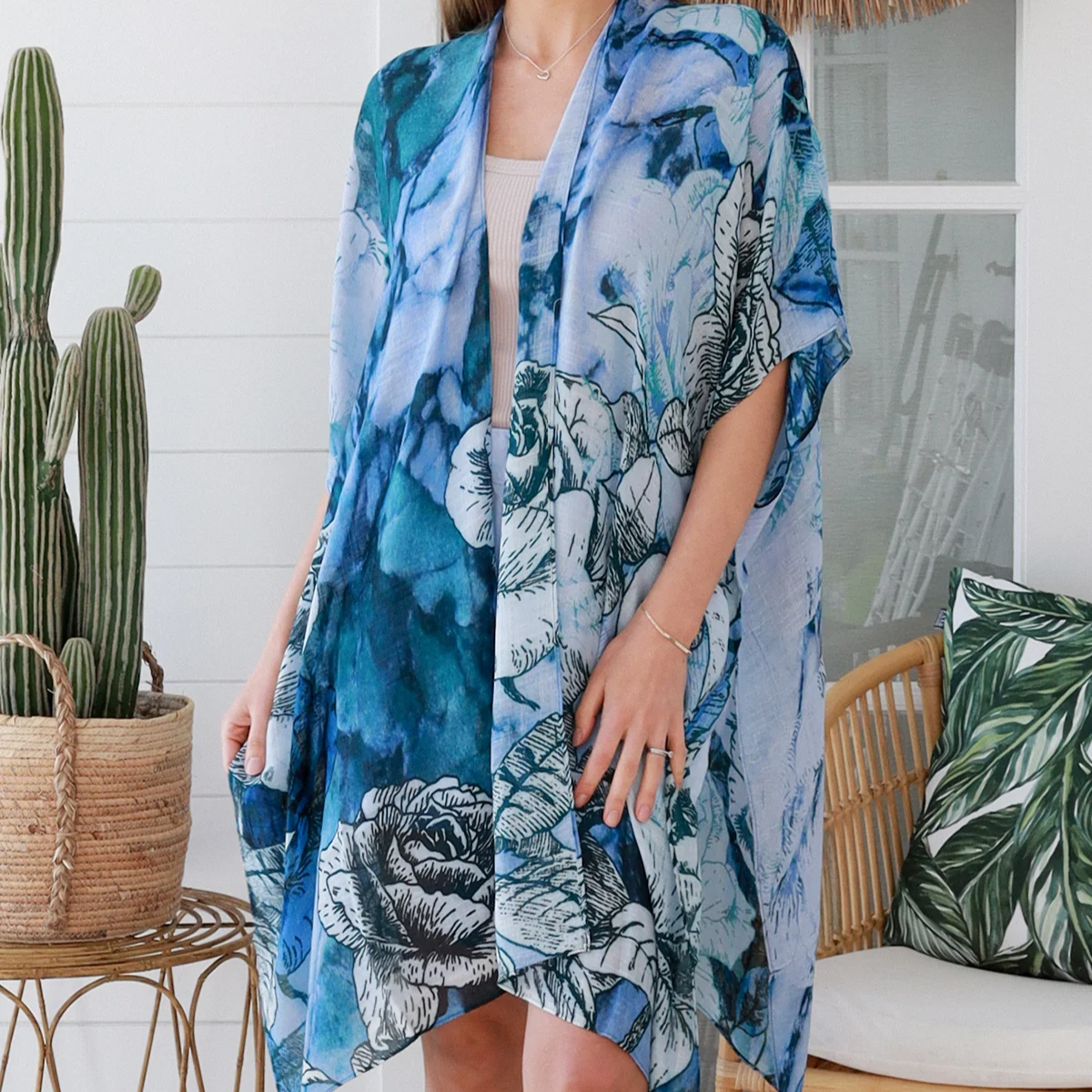 Woman wearing a blue floral kimono standing indoors with a cactus plant and chair in the background.