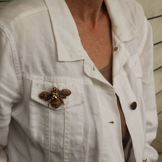 A close-up of a woman wearing a white linen button-up shirt jacket with a decorative brown and gold butterfly brooch pinned to the chest pocket.