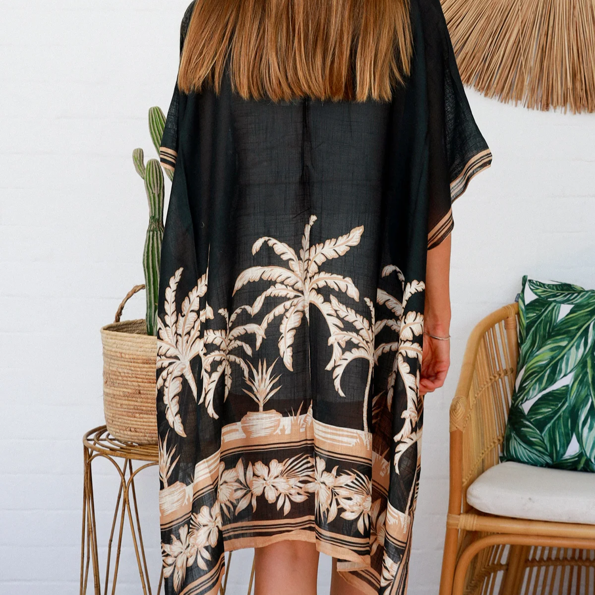 Woman wearing a black floral kimono in a room with a white wall, wicker chair, and decorative items.