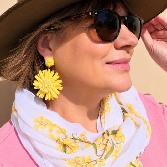 a woman wearing a hat, sunglasses and bright yellow raffia earrings in the shape of a flower and a wattle floral scarf.