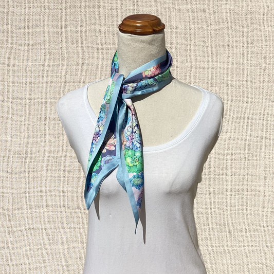A light blue silk scarf with a colorful floral and butterfly print, styled on a cream-colored mannequin torso against a textured beige background.