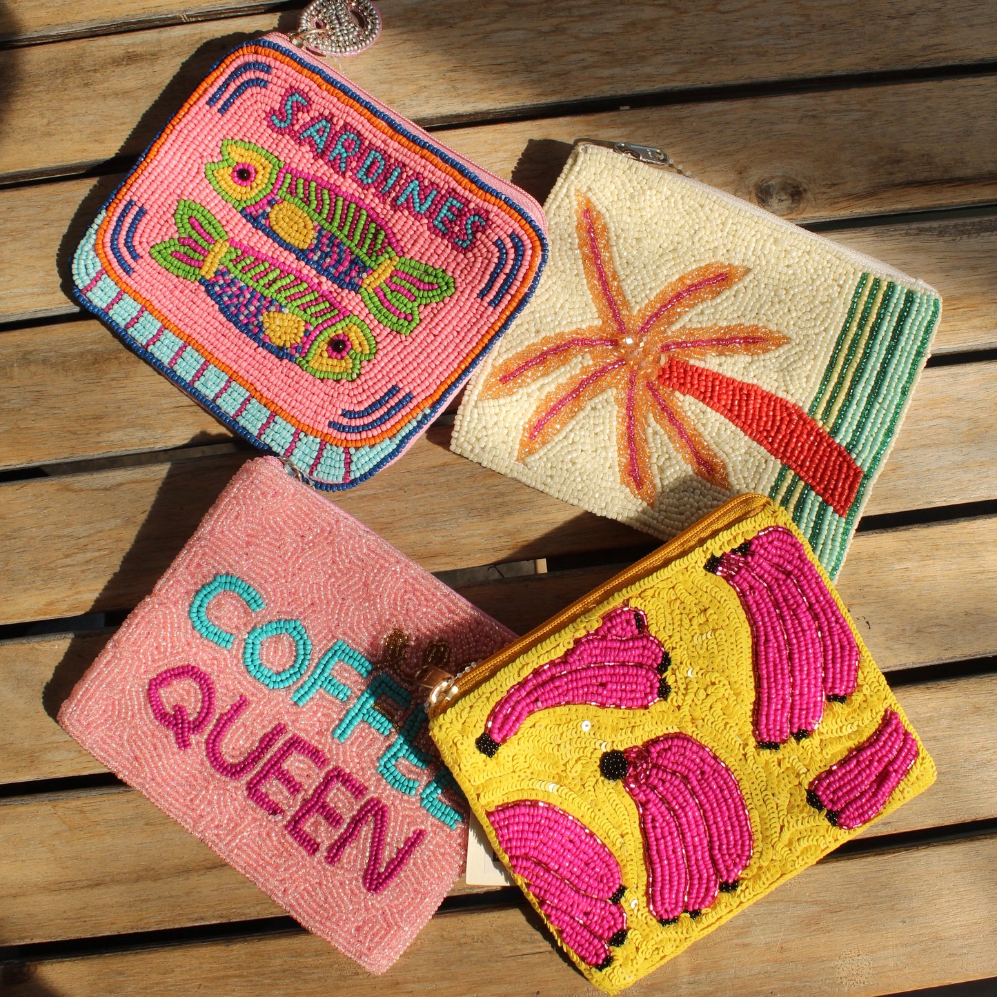 Four colorful beaded zip purses arranged on a wooden slatted surface. The pouches feature vibrant hand-stitched designs including a pink tropical fish design with the word 'SARDINE', a cream palm tree motif, a pink 'COFFEE QUEEN' text design, and a yellow background with pink banana motifs.