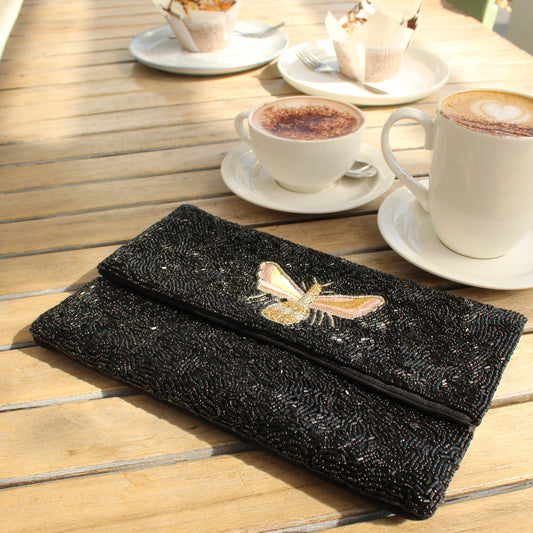 A black textured clutch bag with a bee applique, resting on a wooden cafe table next to coffee cups and pastries.