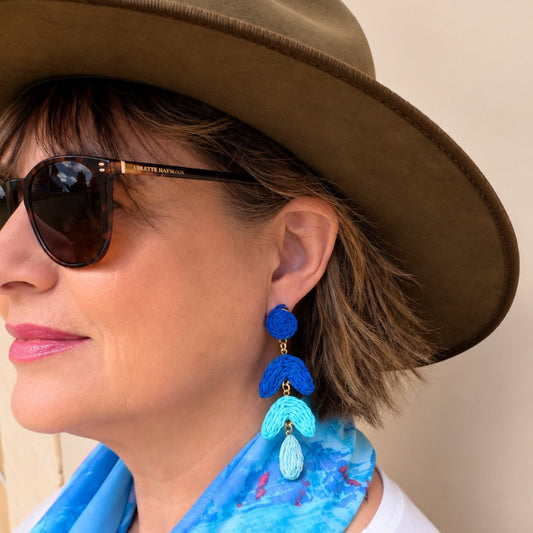 A woman wearing a brown wide-brim hat, sunglasses, a blue patterned scarf, and a white top, shown in profile with a dangling blue and turquoise earring visible.