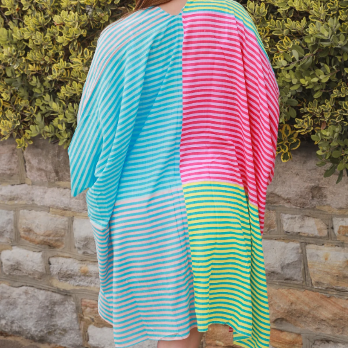 Person wearing a colorful striped kimono standing against a stone wall and green bush.