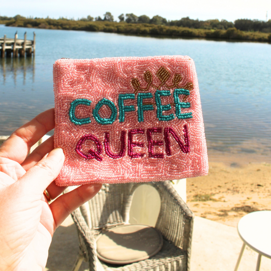 A hand holds a pink beaded square purse with the words 'COFFEE QUEEN' embroidered in teal and magenta, with a small gold crown above. The background shows a lake, dock, and patio chair.