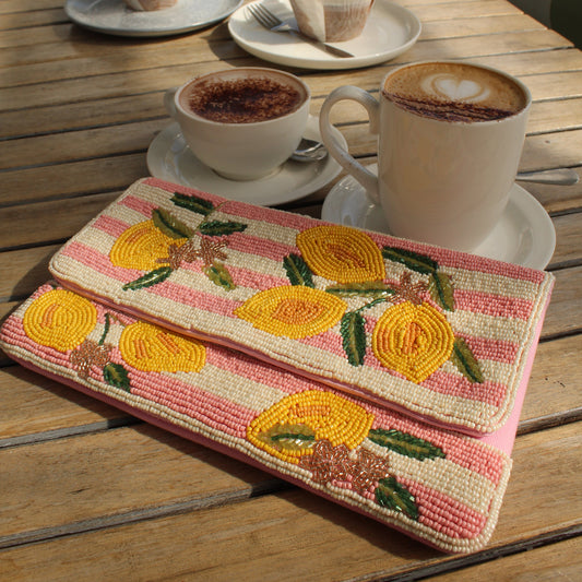 a beaded clutch bag that has a pink and white stripe and lemon pattern on a table with a cups of coffe in the background