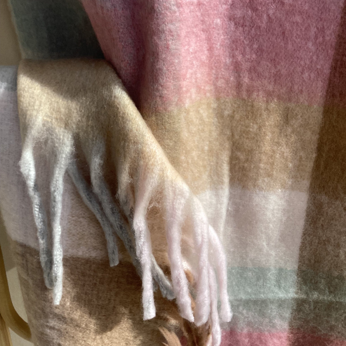 A close-up of a soft plaid sinter scarf with fringe, showing stripes in beige, pink, light gray, and white.
