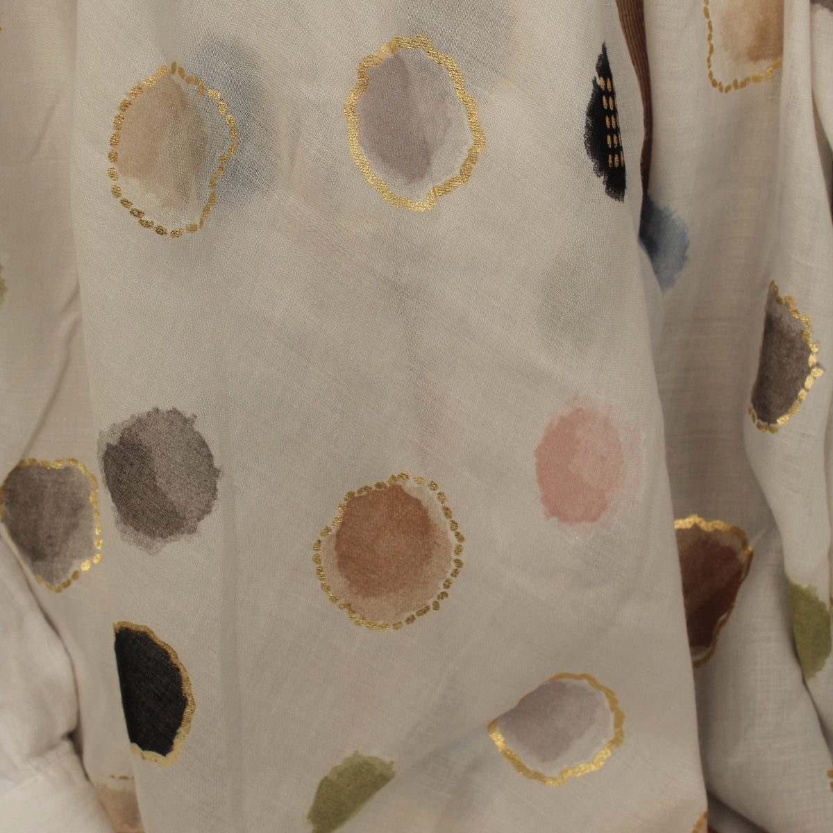 A close-up of a white or off-white fabric with scattered abstract watercolor-style polka dots in muted gray, brown, pink, black, and olive green, each outlined with metallic gold trim.