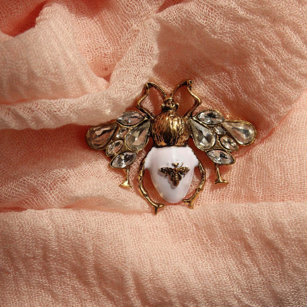 A gold-tone bumblebee brooch with a white enamel center and clear rhinestone wings, resting on draped peach-colored scarf
