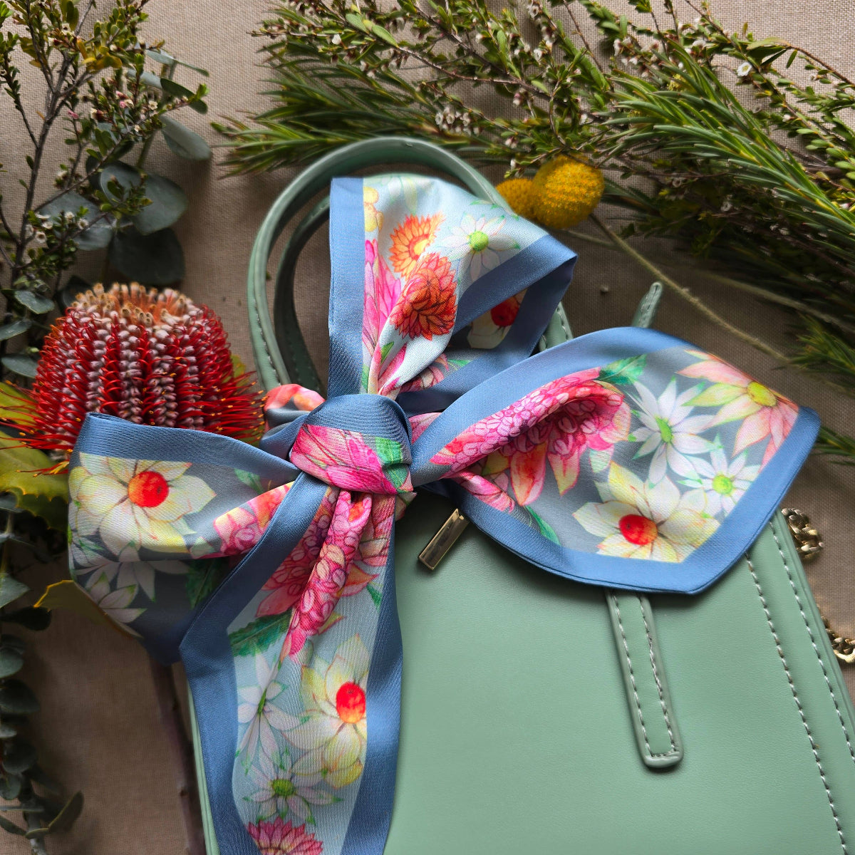 A mint green handbag styled with a large decorative bow made from a floral twilly scarf. The scarf features a light blue border and a vibrant print of pink, red, white, and yellow flowers.