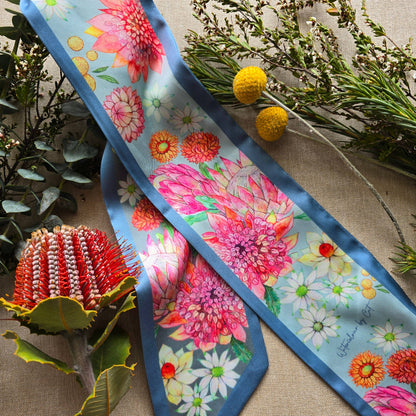 A silky scarf with a vibrant floral print featuring pink protea-like flowers, white daisies, and orange blossoms on a light blue background, styled with natural botanical props on a burlap surface.