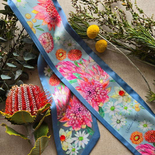 A silky scarf with a vibrant floral print featuring pink protea-like flowers, white daisies, and orange blossoms on a light blue background, styled with natural botanical props on a burlap surface.