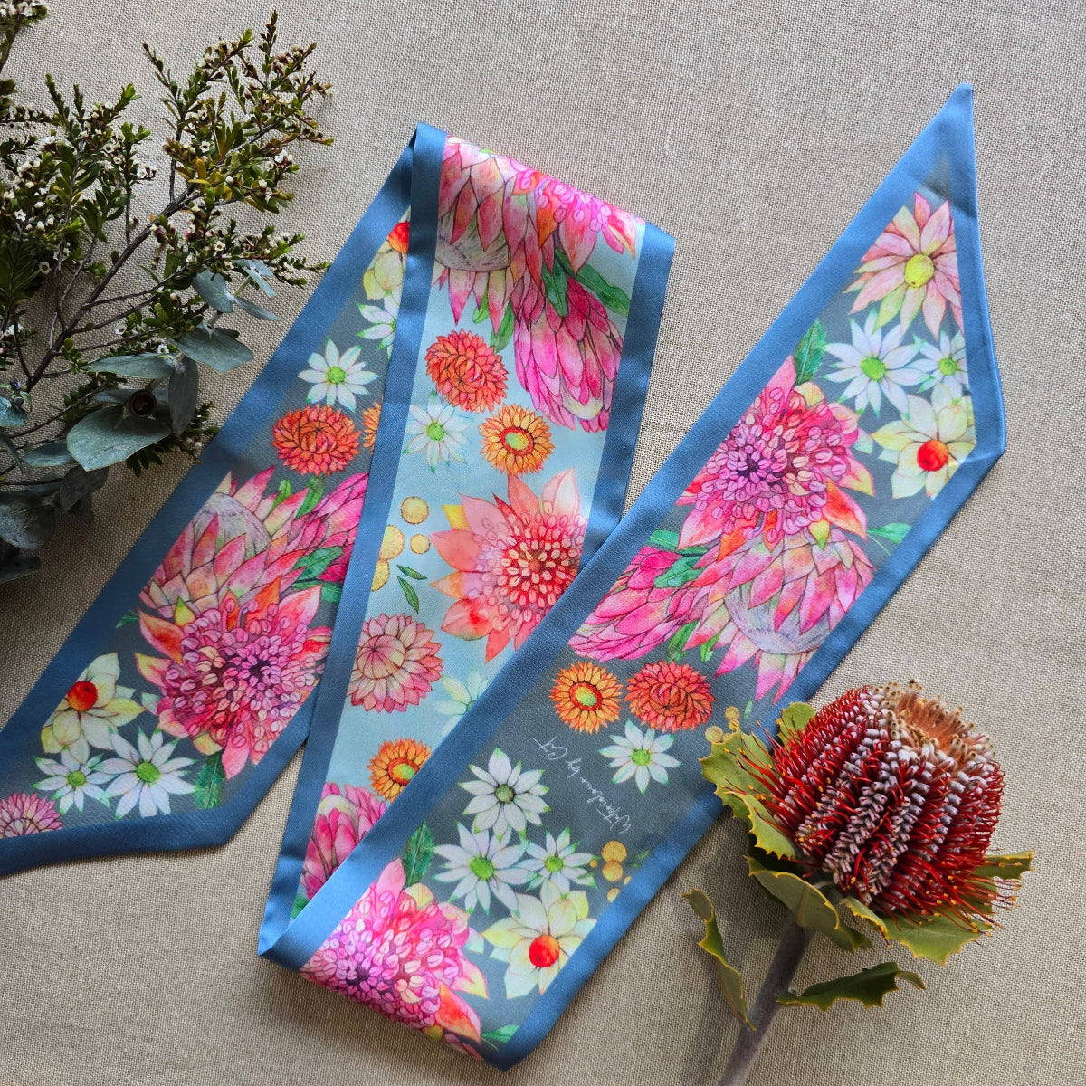 A silky scarf with a vibrant floral print featuring pink, orange, and white flowers on a light blue background with a medium blue border, styled on a beige linen fabric with decorative greenery and a protea flower.