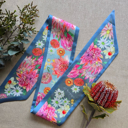 A silky scarf with a vibrant floral print featuring pink, orange, and white flowers on a light blue background with a medium blue border, styled on a beige linen fabric with decorative greenery and a protea flower.