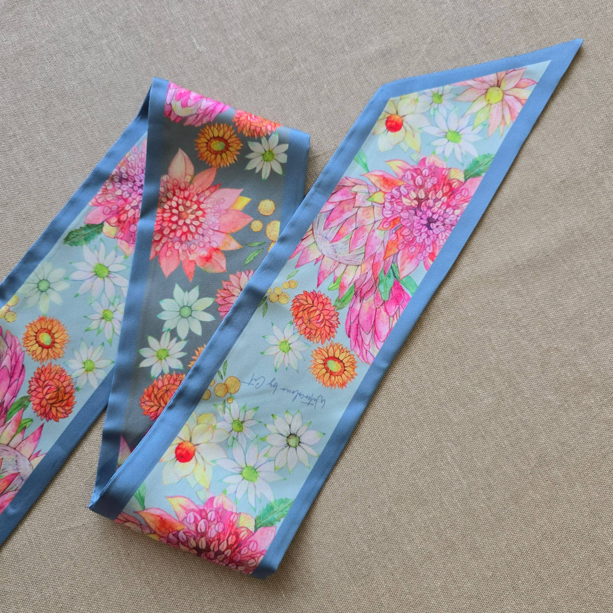 A floral silky scarf with a blue border, featuring a vibrant print of pink, orange, and white flowers on a light blue background, laid out on a beige textured fabric surface.