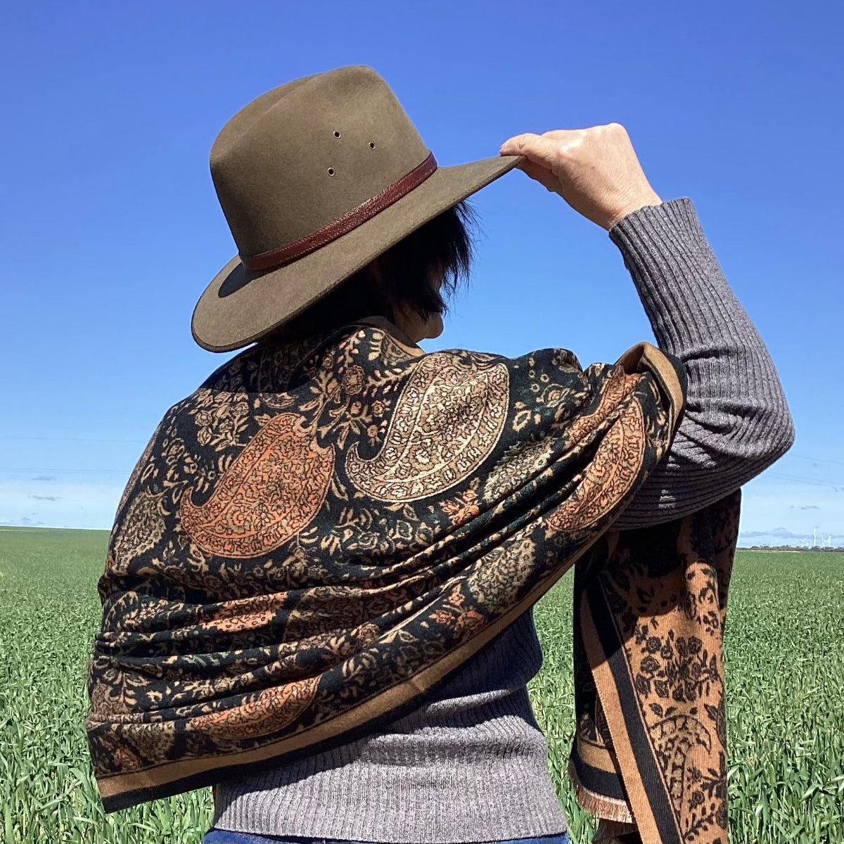 Person wearing a patterned shawl and hat in an open field with blue sky.