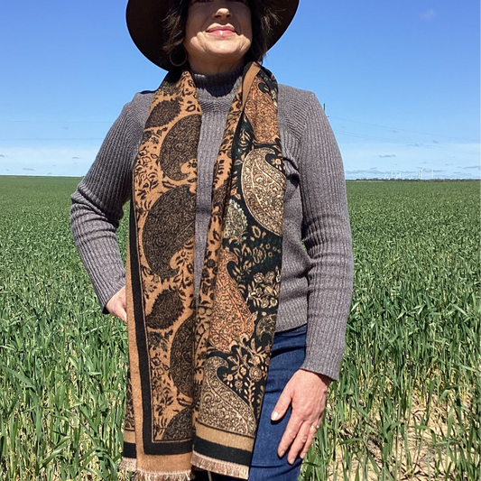 A lady wearing a patterned scarf, hat, and gray sweater standing in a field with a blue sky.
