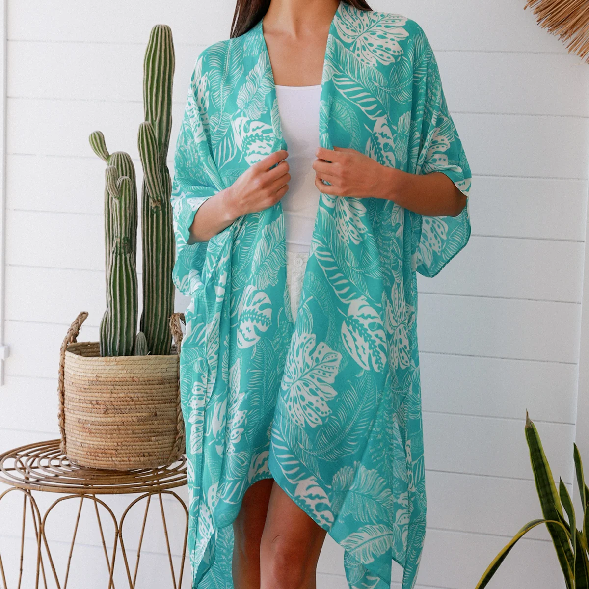 Woman wearing a teal floral kimono in a room with plants and a white wall.