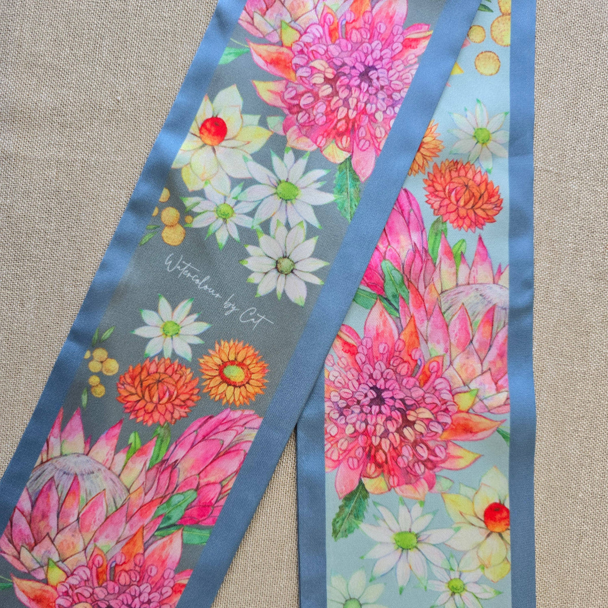 A floral silky scarf laid flat on a textured beige fabric background. The scarf features a vibrant watercolor-style floral print with pink protea-like flowers, white daisies, and orange ditsy blooms on a dusty blue-gray background, framed by solid medium-blue borders.
