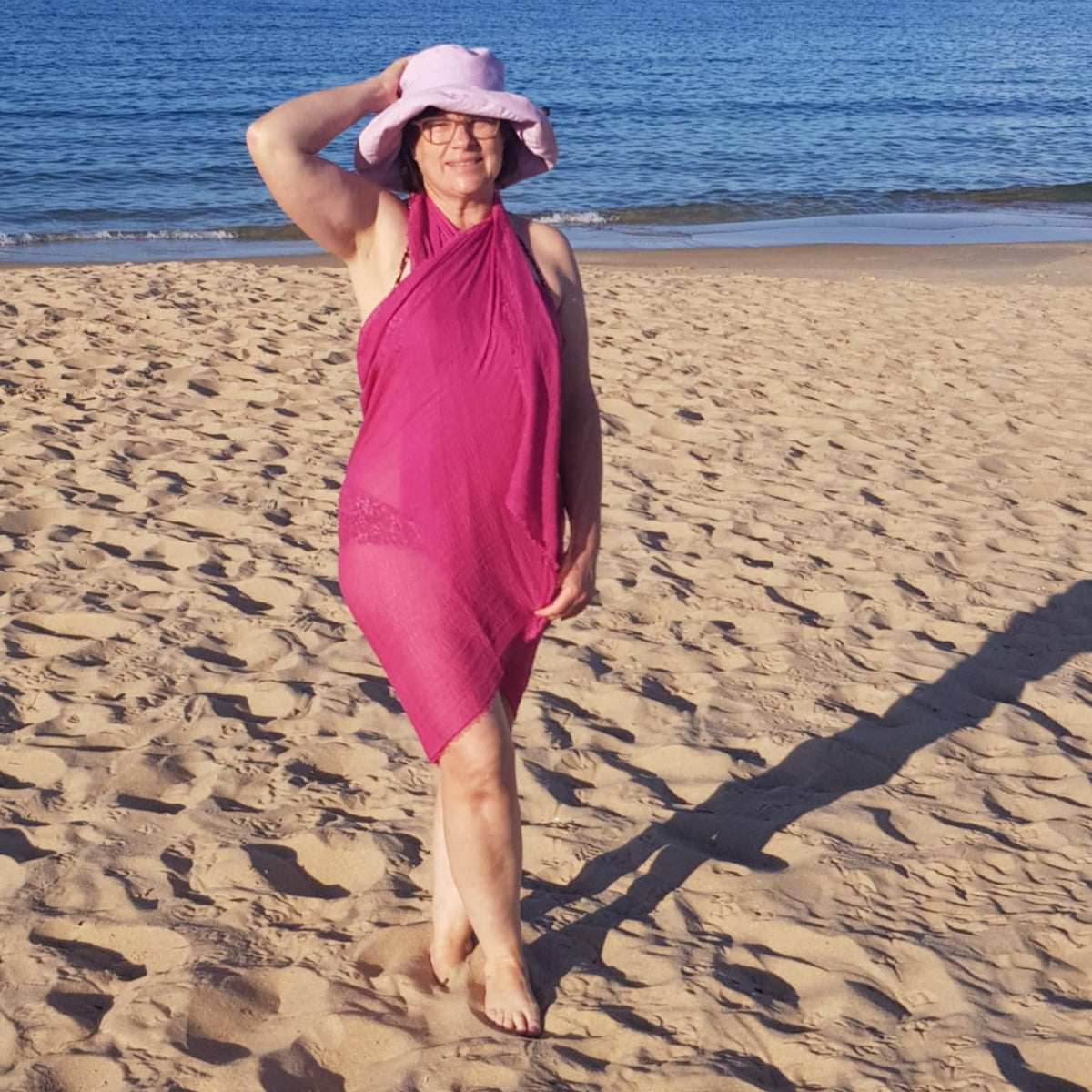 Woman in a pink scarf that is draped as a sarong and wearing a light pink hat standing on a sandy beach with blue water in the background