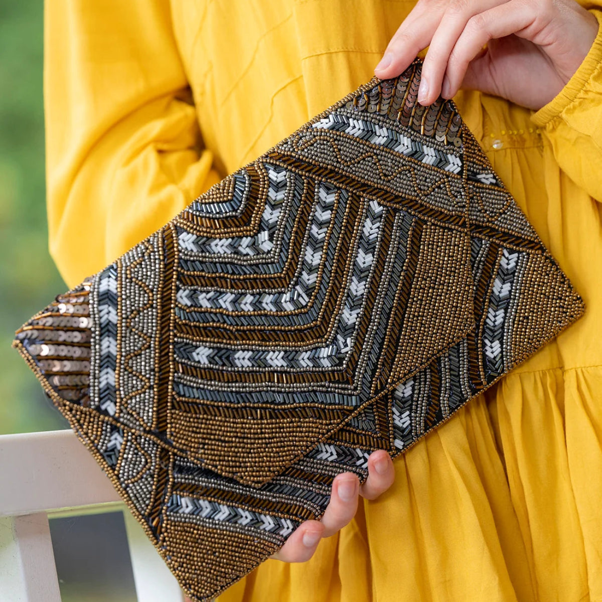 A person in a bright yellow dress holds a structured envelope-style clutch bag decorated with intricate geometric beadwork in brown, black, and white.