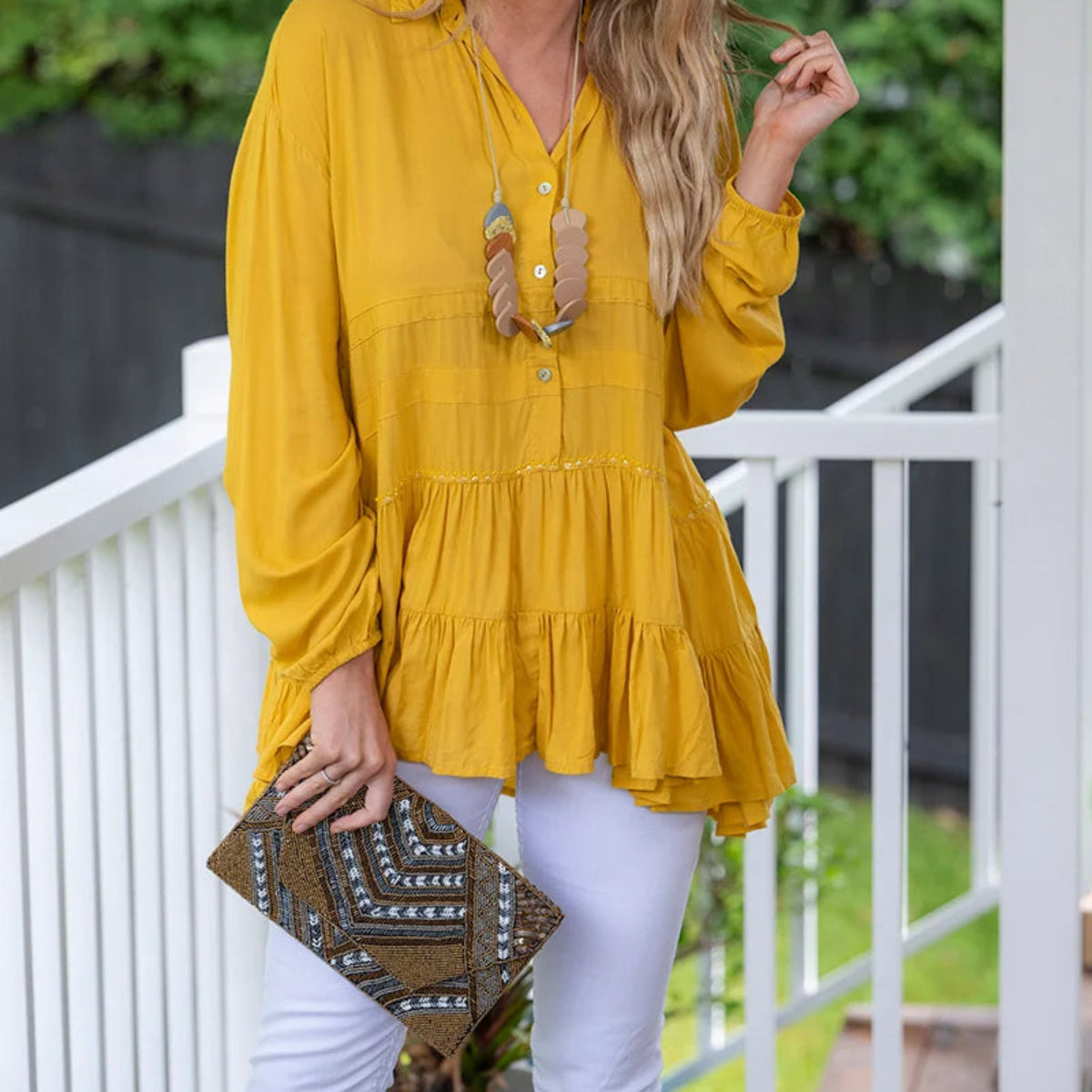 A woman stands on a white porch wearing a mustard yellow long-sleeve tiered blouse and white pants, holding a brown patterned clutch. The background features green foliage and a white railing.