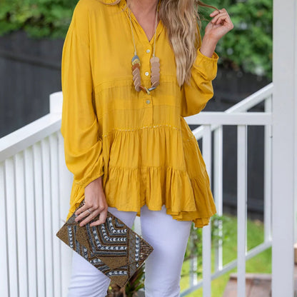 A woman stands on a white porch wearing a mustard yellow long-sleeve tiered blouse and white pants, holding a brown patterned clutch. The background features green foliage and a white railing.