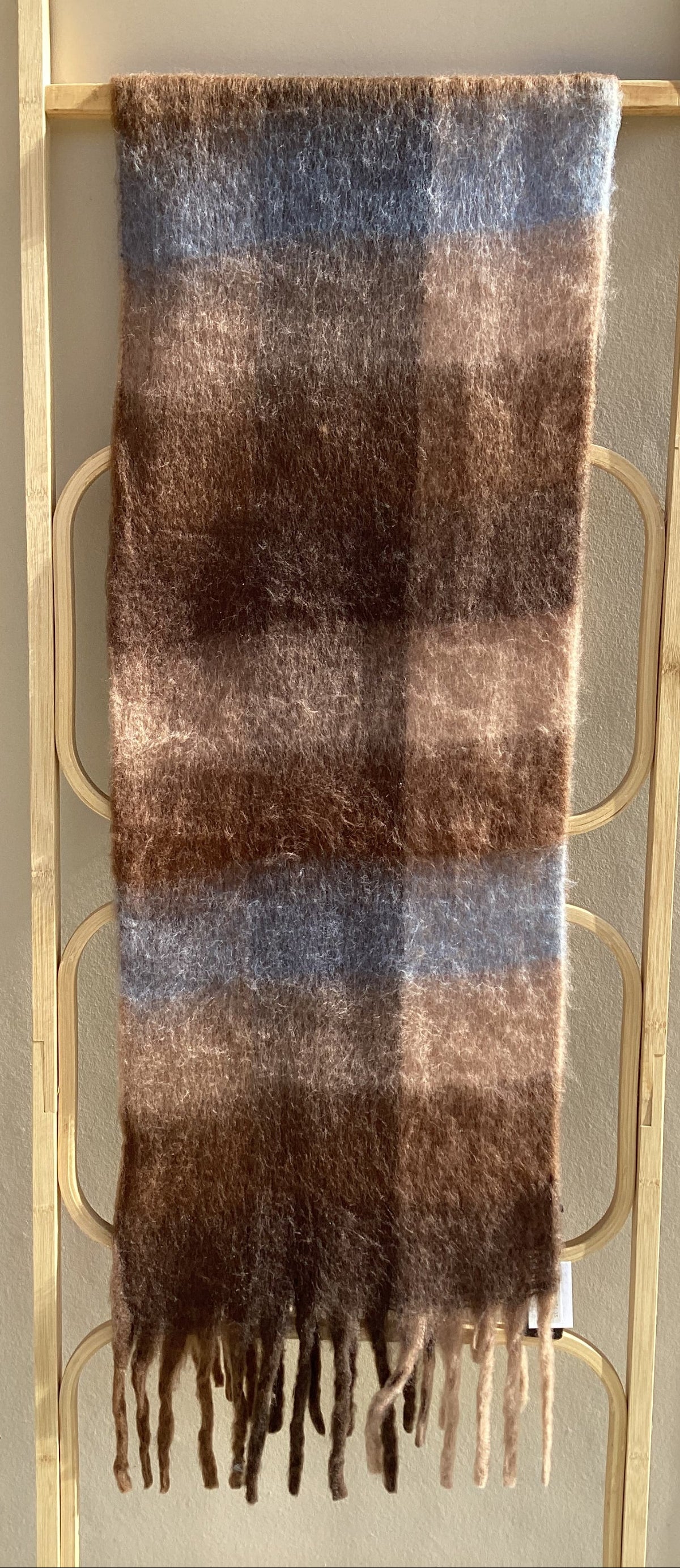 A soft, fluffy plaid scarf in brown, beige, and muted blue tones, draped over a wooden rack against a neutral beige wall.