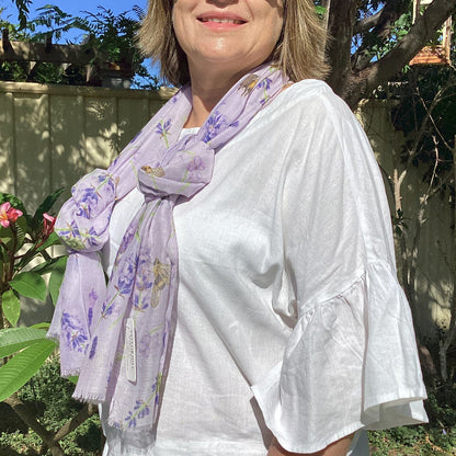 Woman wearing sunglasses and a white outfit with a lilac purple floral scarf outdoors.