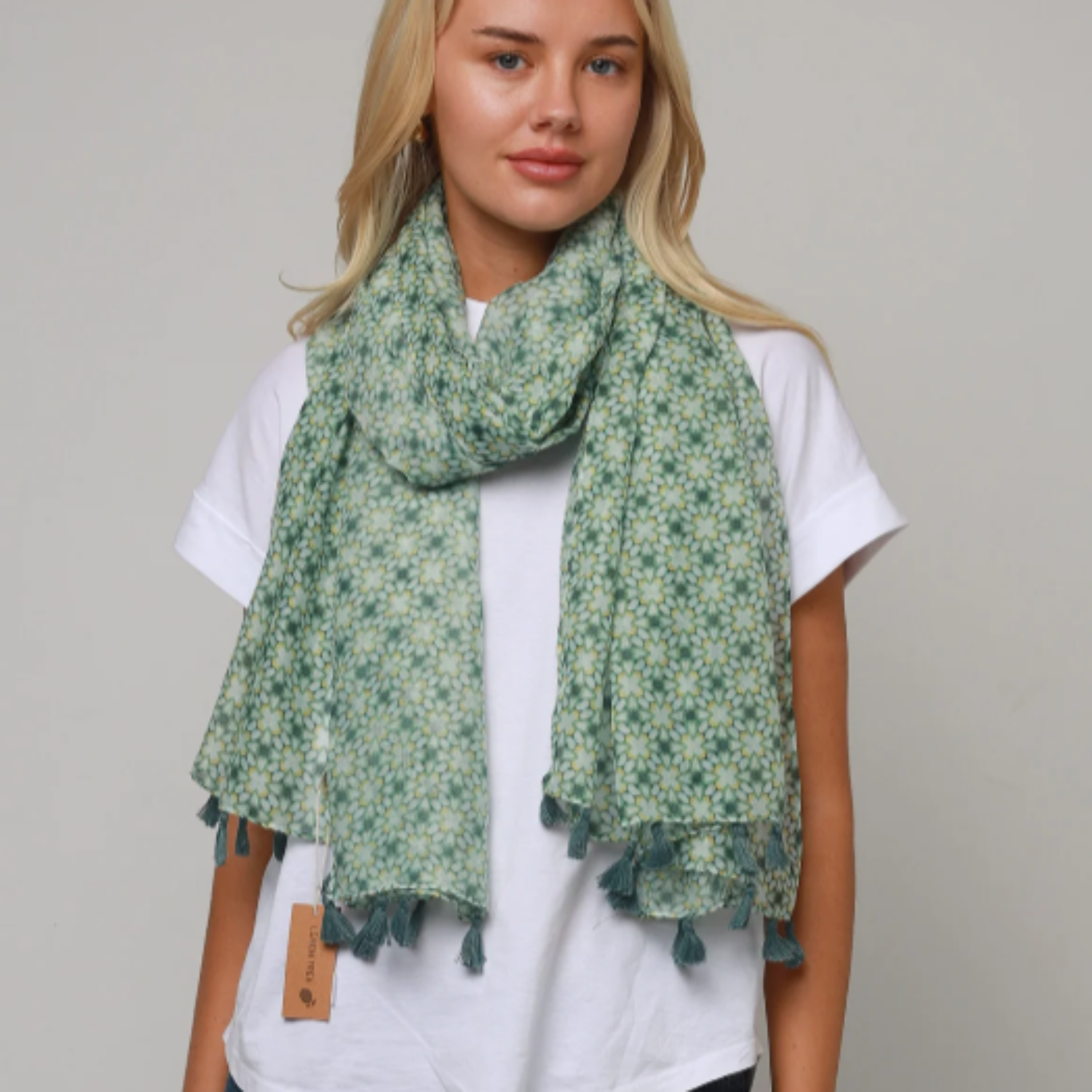 a lady wearing a scarf that has an small abstract floral pattern in colours of sage green,pale marigold, white and finished with dark green tassels.