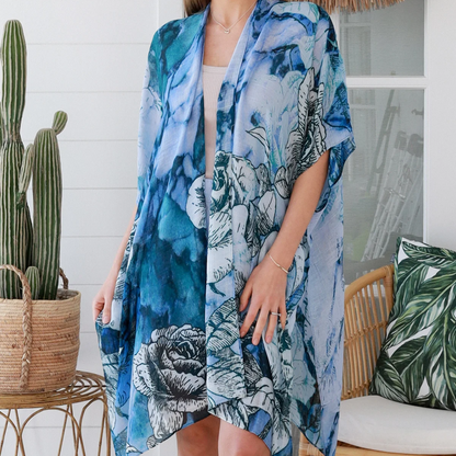 Woman wearing a blue floral kimono standing indoors with a cactus plant and chair in the background.