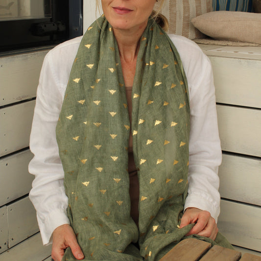 A woman wearing a sage green scarf with small gold bee motifs, styled with a white shirt and sunglasses, sitting on a wooden bench.