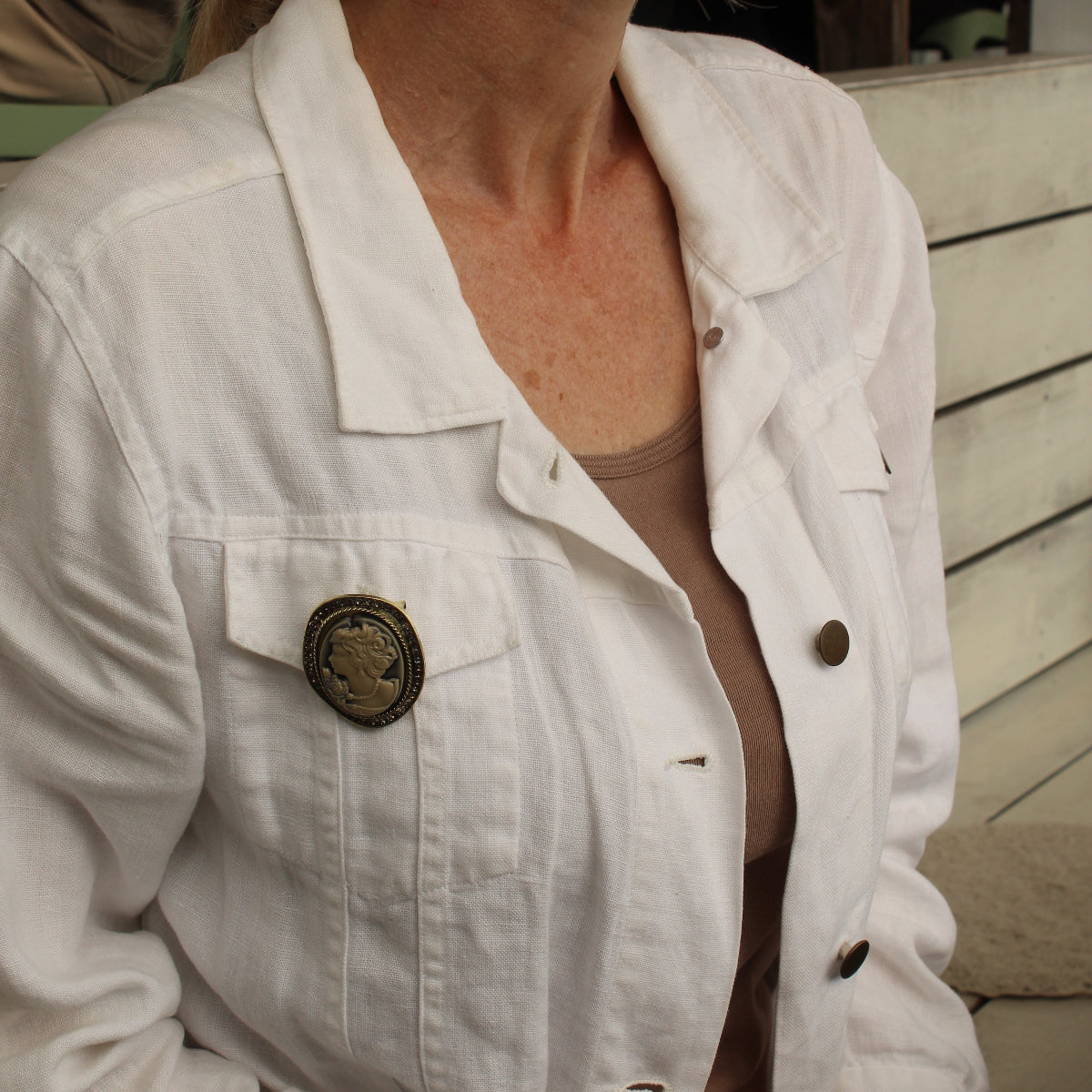 A woman wearing a white denim jacket with a prominent round bronze button on the chest pocket, layered over a beige top.