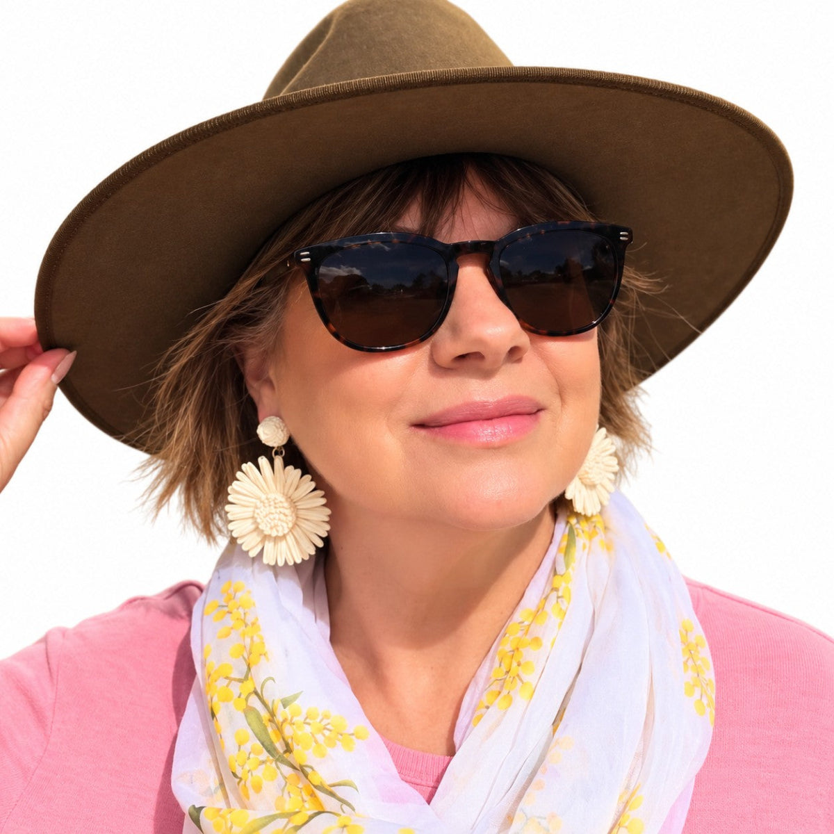 A woman wearing a wide-brimmed brown hat, dark sunglasses, large cream flower-shaped earrings, and a white scarf with yellow floral print over a pink top, photographed against a plain white background.