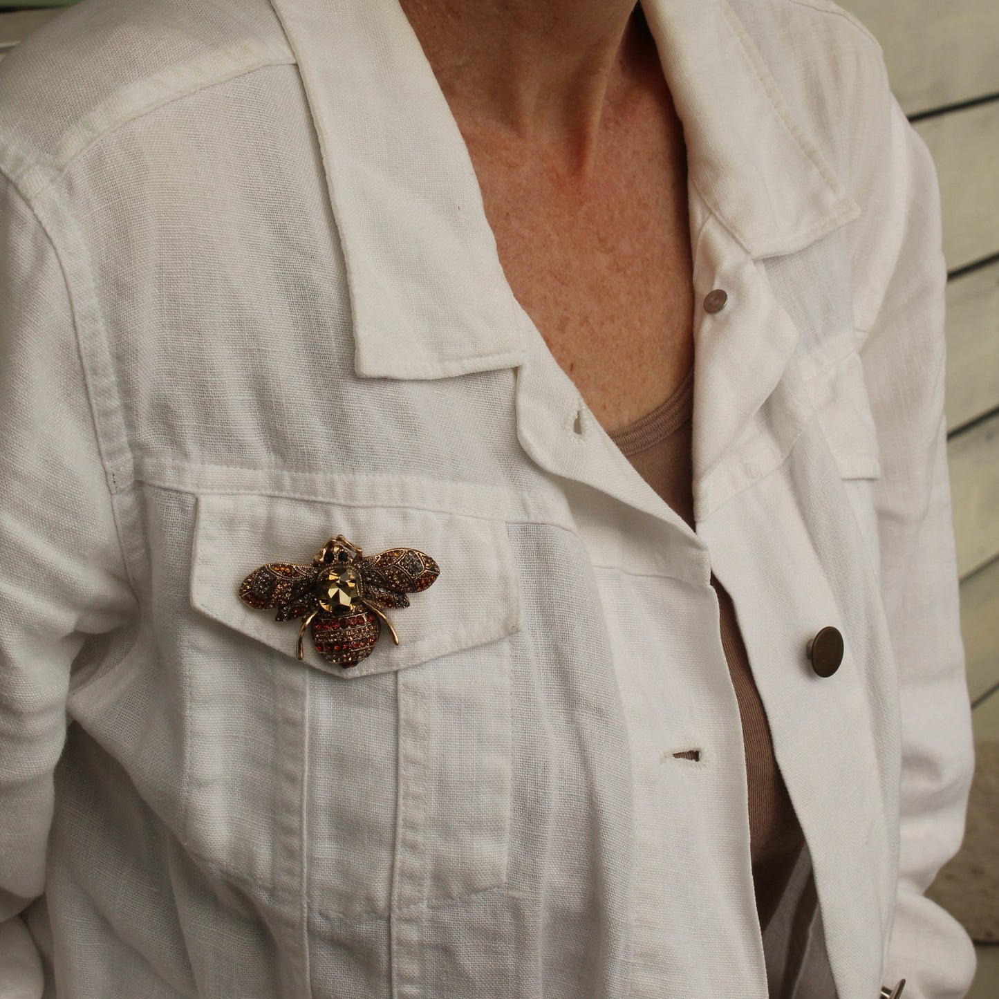 A close-up of a woman wearing a white linen button-up shirt jacket with a decorative brown and gold butterfly brooch pinned to the chest pocket.