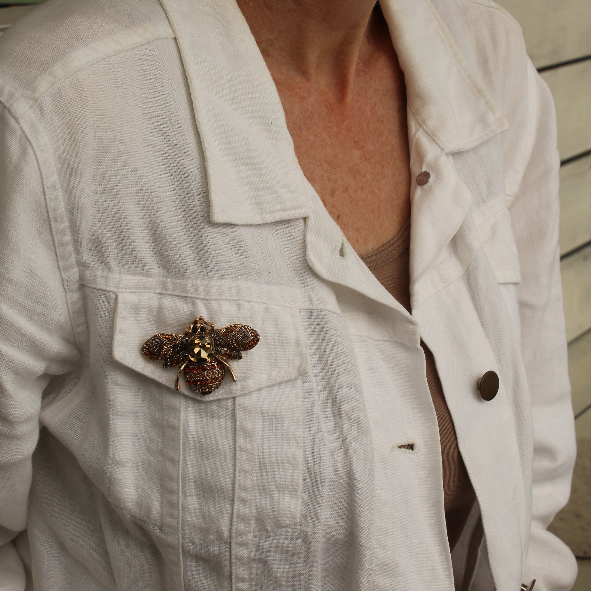 A close-up of a woman wearing a white linen button-up shirt jacket with a decorative brown and gold butterfly brooch pinned to the chest pocket.