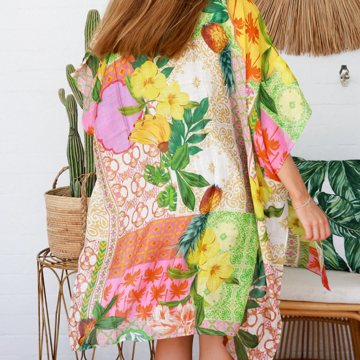 Woman wearing a colorful floral kimono in a room with a cactus and woven chair.