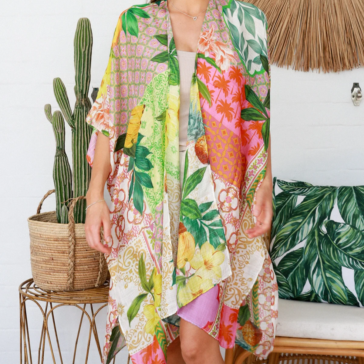 Woman wearing a colorful floral kimono in a tropical setting with plants and a woven wall hanging.