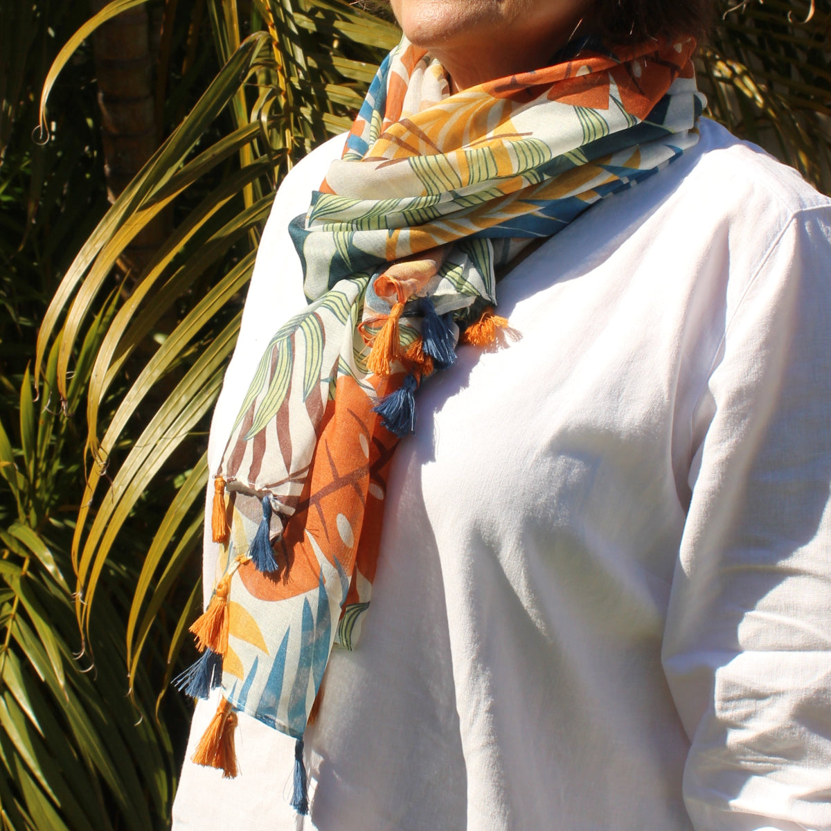 Person wearing a tropical patterned scarf and white shirt standing among palm leaves