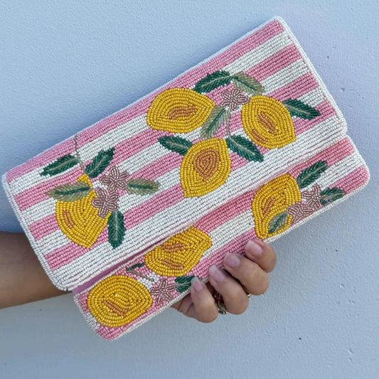 A hand holding a rectangular beaded clutch purse with a pink and white striped background and yellow lemon motifs, set against a light blue wall.