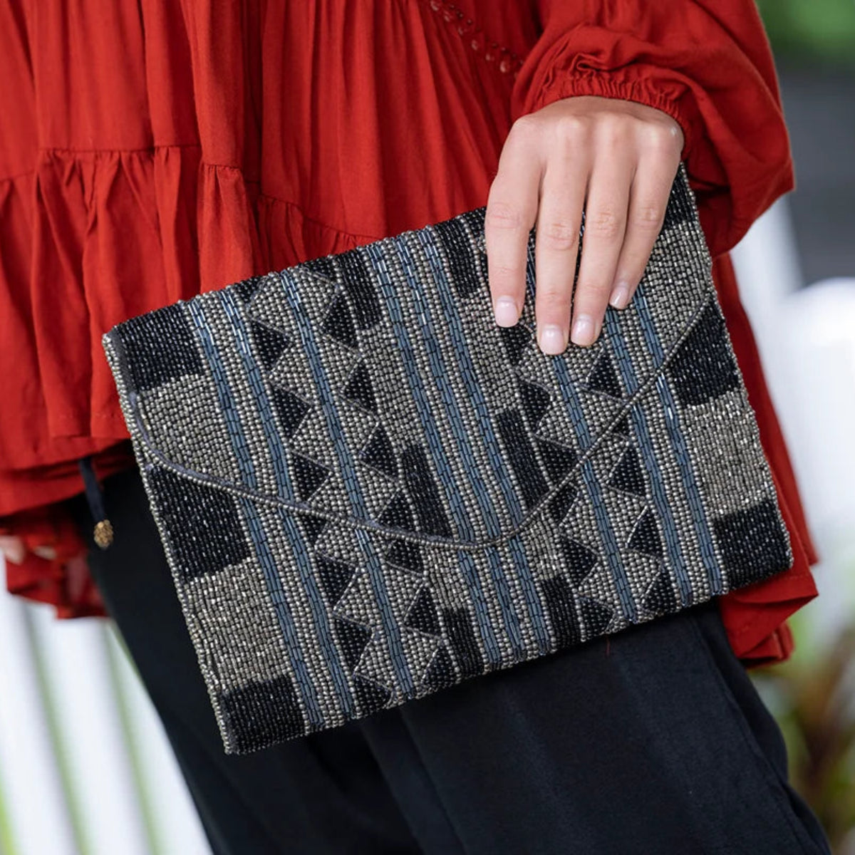 A person in a red blouse holds a woven envelope-style clutch with a geometric pattern in black, gray, beige, and muted blue. The background is a softly blurred outdoor setting with white railings.