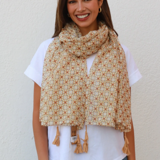 a lady wearing a scarf that has an abstract floral pattern in shades of caramel brown, muted orange, grey and finished with caramel brown tassles