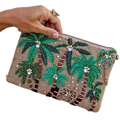A hand holds a small zip pouch decorated with embroidered palm trees and small pearl-like beads on a tan woven background, against a plain light wall.